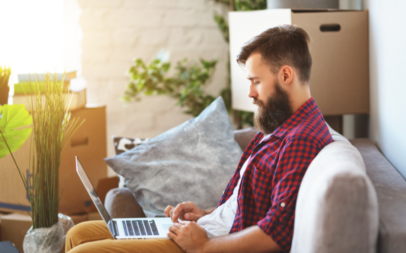 Bearded man in a red flannel using his laptop and surrounded by moving boxes
